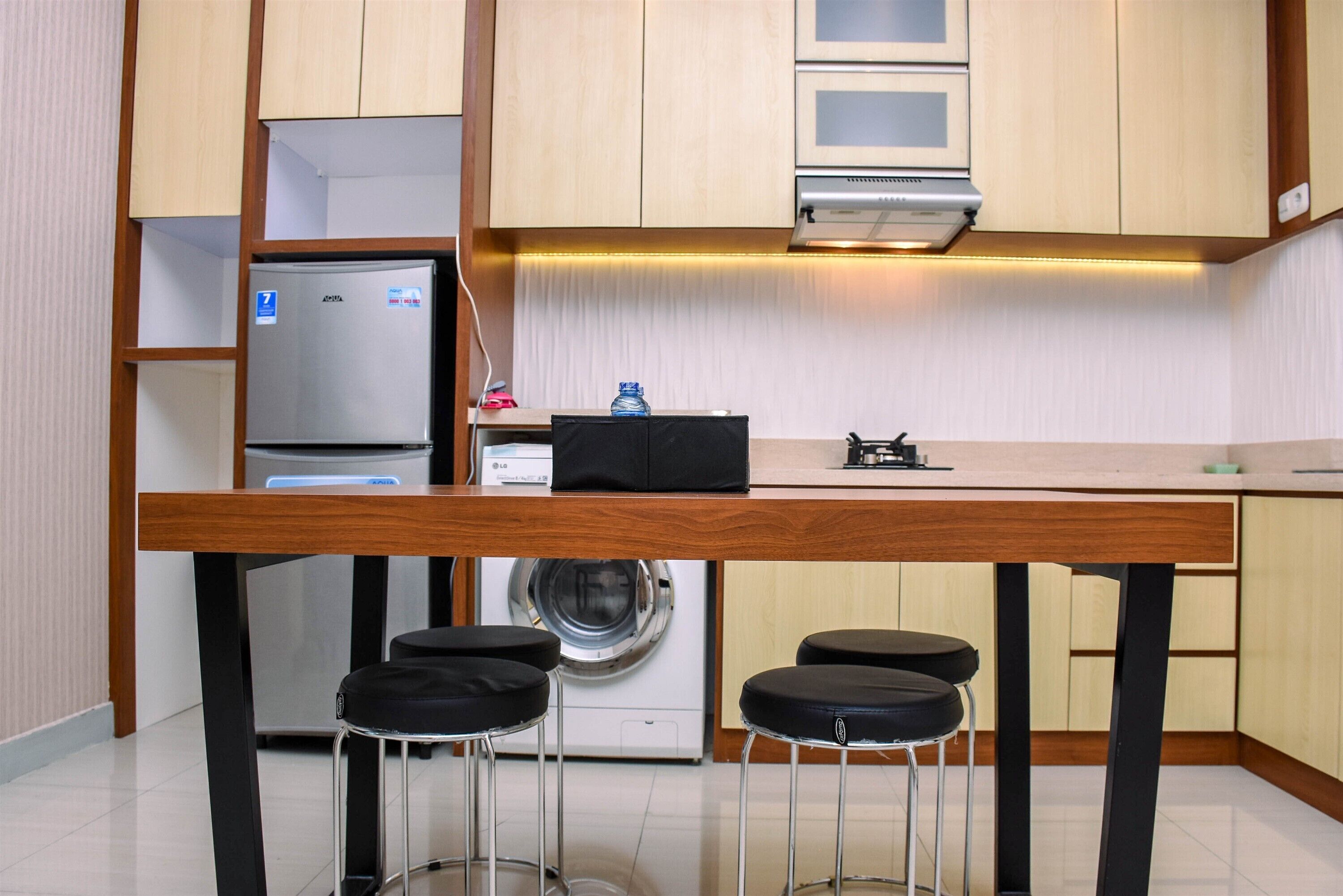 A compact kitchen with a built-in refrigerator, microwave, and washing machine. There are three stools and a small table.