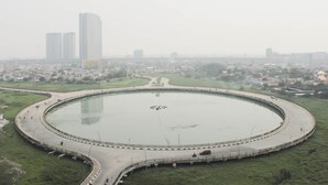 Een groot, rond waterlichaam met een omliggende weg, omgeven door groen en in de verte gebouwen.