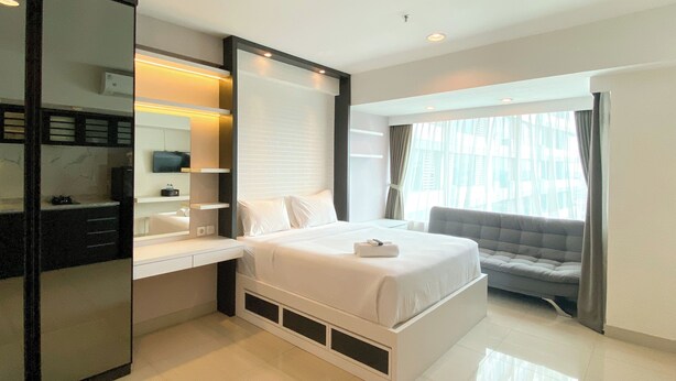A modern hotel room with a large bed, a built-in desk, a vanity with a mirror, and a window with curtains. - Well Furnished And Cozy Stay Studio Grand Kamala Lagoon Apartment (Bekasi)