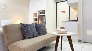 A modern hotel room with a beige sofa, patterned cushions, a small round table, and a kitchenette area. - Minimalist And Homey Studio At Grand Kamala Lagoon Apartment (Bekasi)