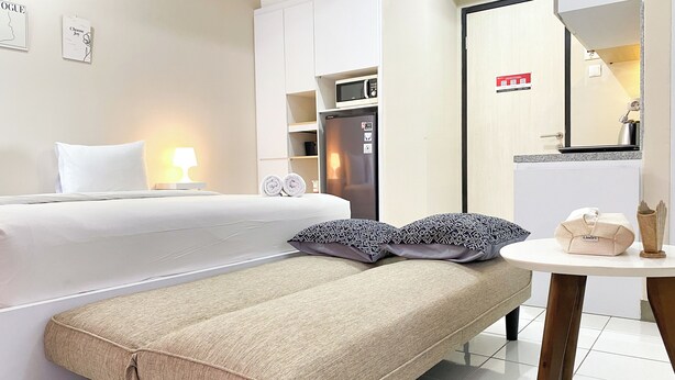 A hotel room with a bed, two pillows, a bedside table with a lamp, a microwave, and a refrigerator. - Minimalist And Homey Studio At Grand Kamala Lagoon Apartment (Bekasi)