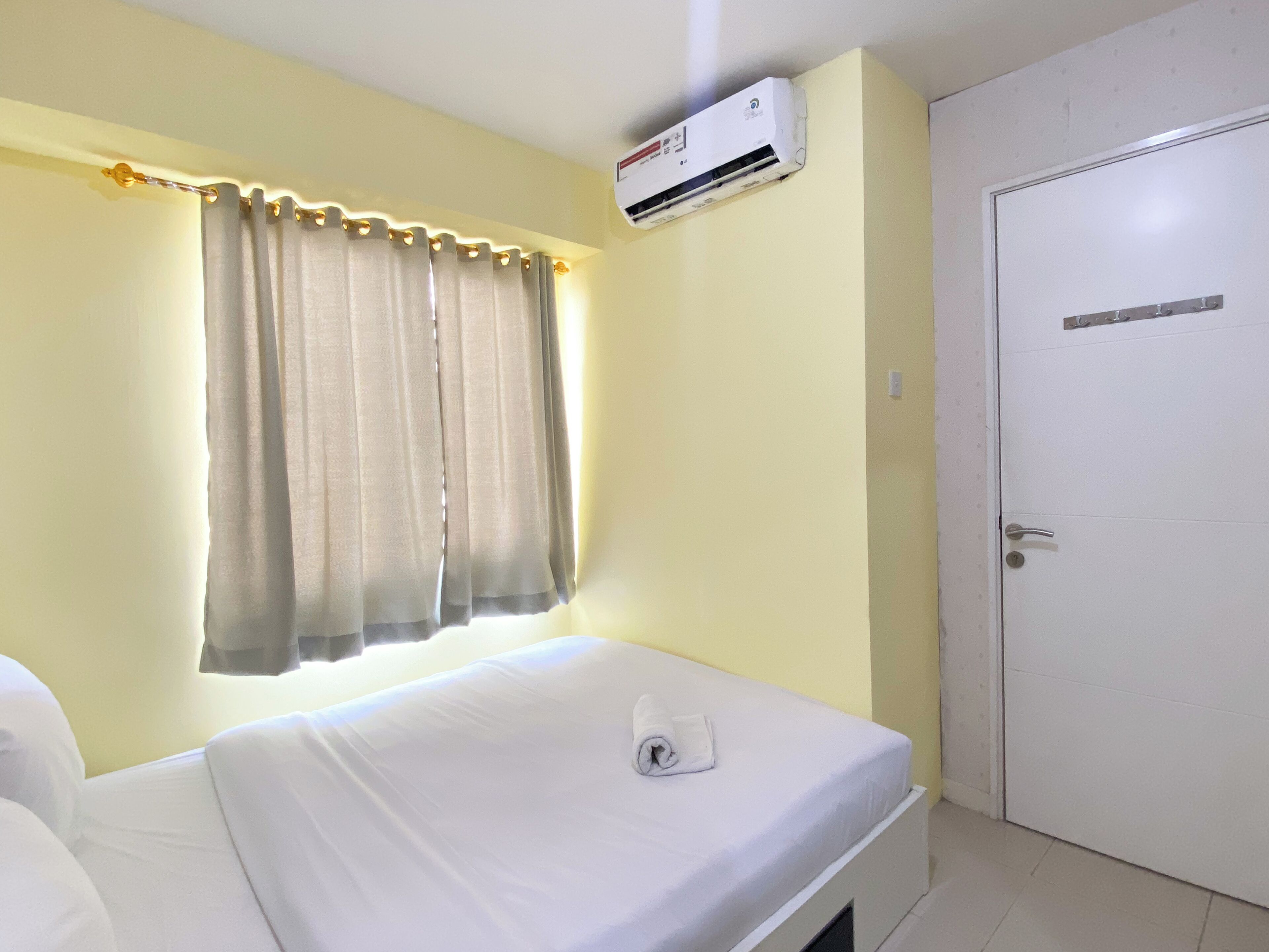 A single-bed room with a white mattress, a window with curtains, an air conditioning unit, and a white door.