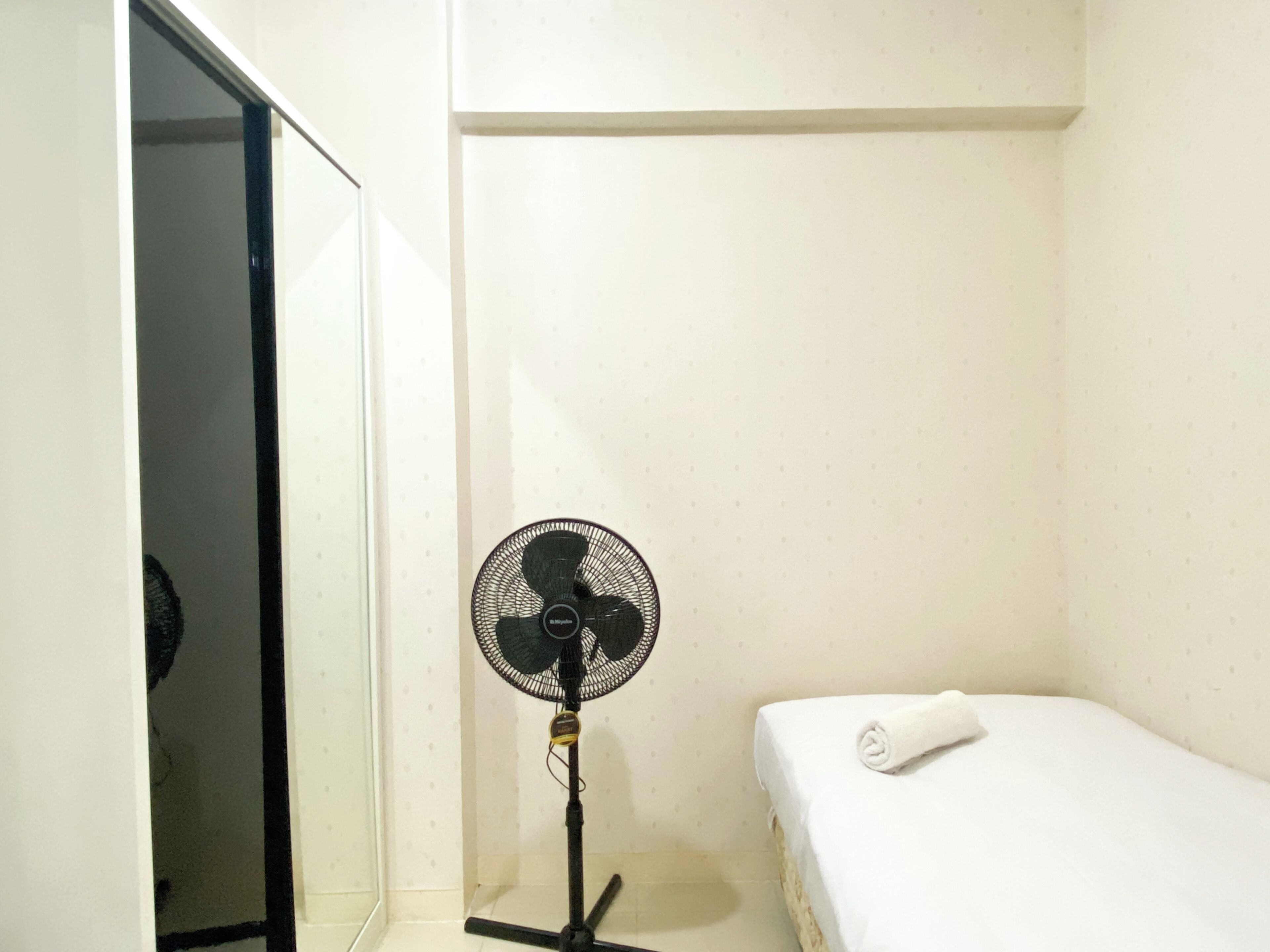 A small, single-bed room with a standing fan and a mirror on the left wall.