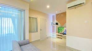 A modern living room with a grey sofa, a flat-screen TV, and a large window with sheer curtains. - Homey And Well Furnished 2Br Grand Kamala Lagoon Apartment (Bekasi)