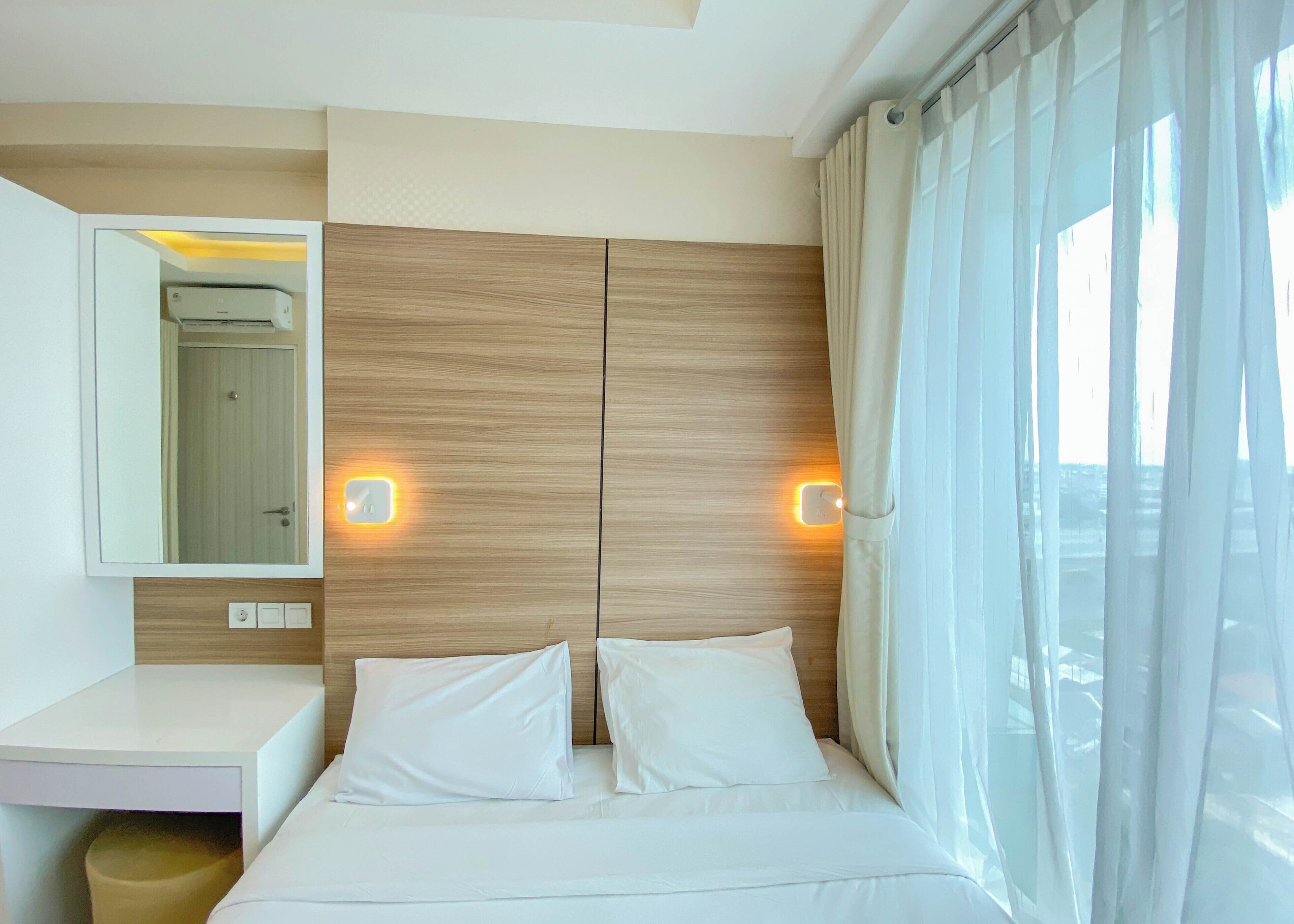 Una habitación de hotel con cabecera de madera, un espejo grande, dos lámparas de noche, una cama con ropa blanca y una ventana con cortinas translúcidas.