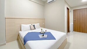 A hotel room with a bed, a blue throw blanket, a small table with a vase, and an air conditioning unit on the wall. - Good And Spacious Studio Lrt City Royal Sentul Park Apartment (Bogor)