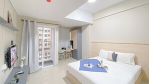 A hotel room with a bed, a desk, a chair, a TV, and a view of the outside. - Good And Spacious Studio Lrt City Royal Sentul Park Apartment (Bogor)