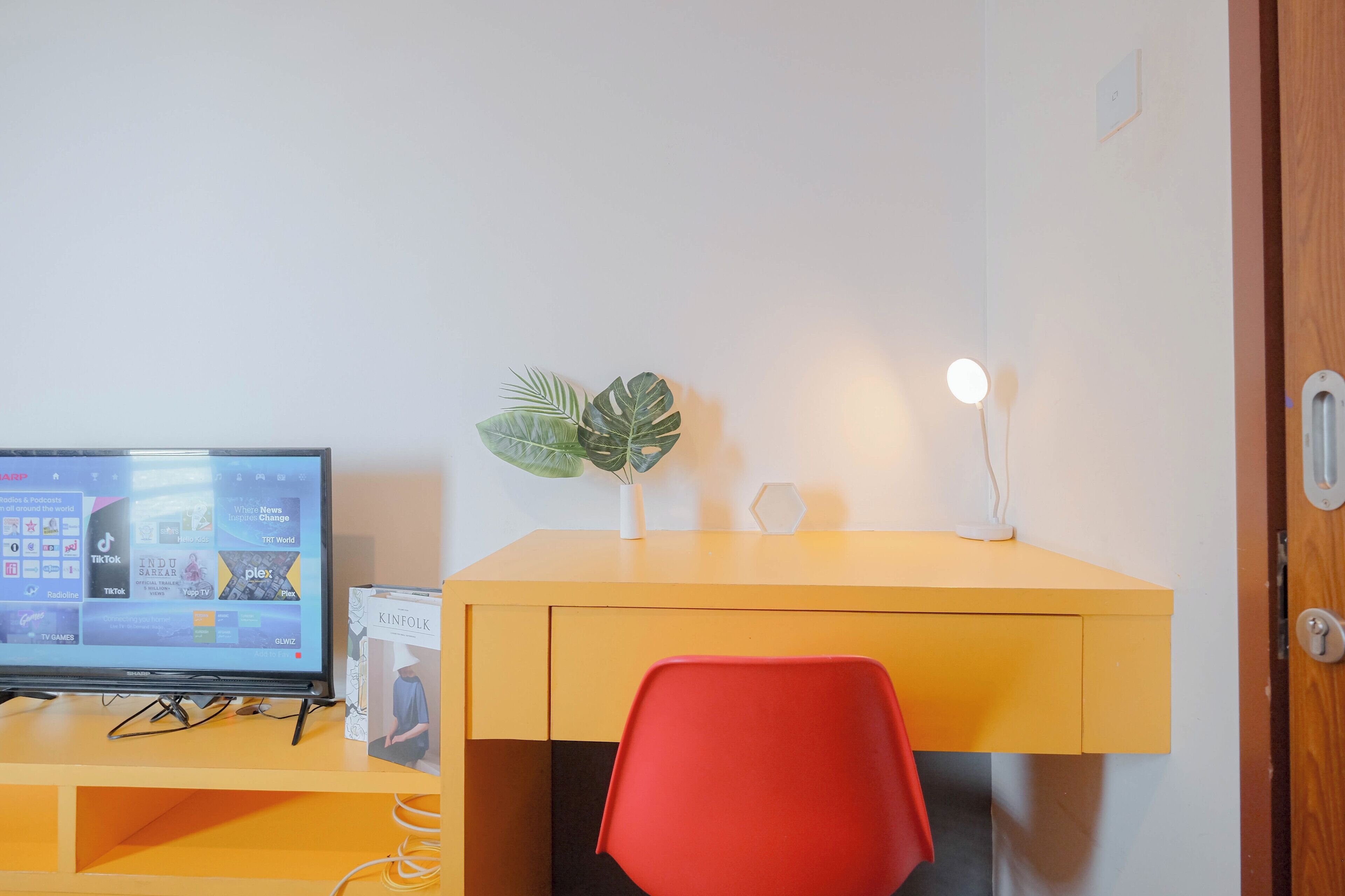 A room with a desk, a red chair, a television, a plant, and a wall-mounted light.
