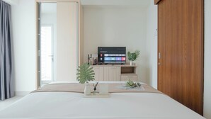 A modern bedroom with a bed, a TV on a stand, a wooden wardrobe, and a sliding door. - Serene Living Studio Apartment Grand Kamala Lagoon (Bekasi)
