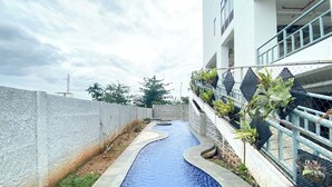 A swimming pool with a curved design, bordered by a concrete wall and a building with balconies. - Compact Wood Accents Studio Apartment Grand Kamala Lagoon (Bekasi)