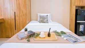A small, neatly arranged bedroom with a bed, a bedside table with a plant, books, a candle, and a tray with a cup and saucer. - Compact Wood Accents Studio Apartment Grand Kamala Lagoon (Bekasi)