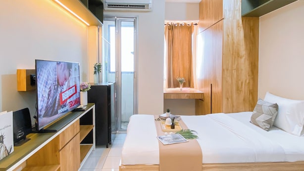 A hotel room with a bed, a TV, a desk, and a window with curtains. - Compact Wood Accents Studio Apartment Grand Kamala Lagoon (Bekasi)