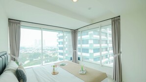 A modern hotel room with a large bed, a bedside table with a lamp, and a view of the city through floor-to-ceiling windows. - Modern Peach Touch 2Br At Grand Kamala Lagoon Apartment (Bekasi)