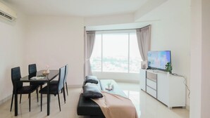 A modern living room with a sofa, a dining table with chairs, a TV, and a large window with curtains. - Modern Peach Touch 2Br At Grand Kamala Lagoon Apartment (Bekasi)