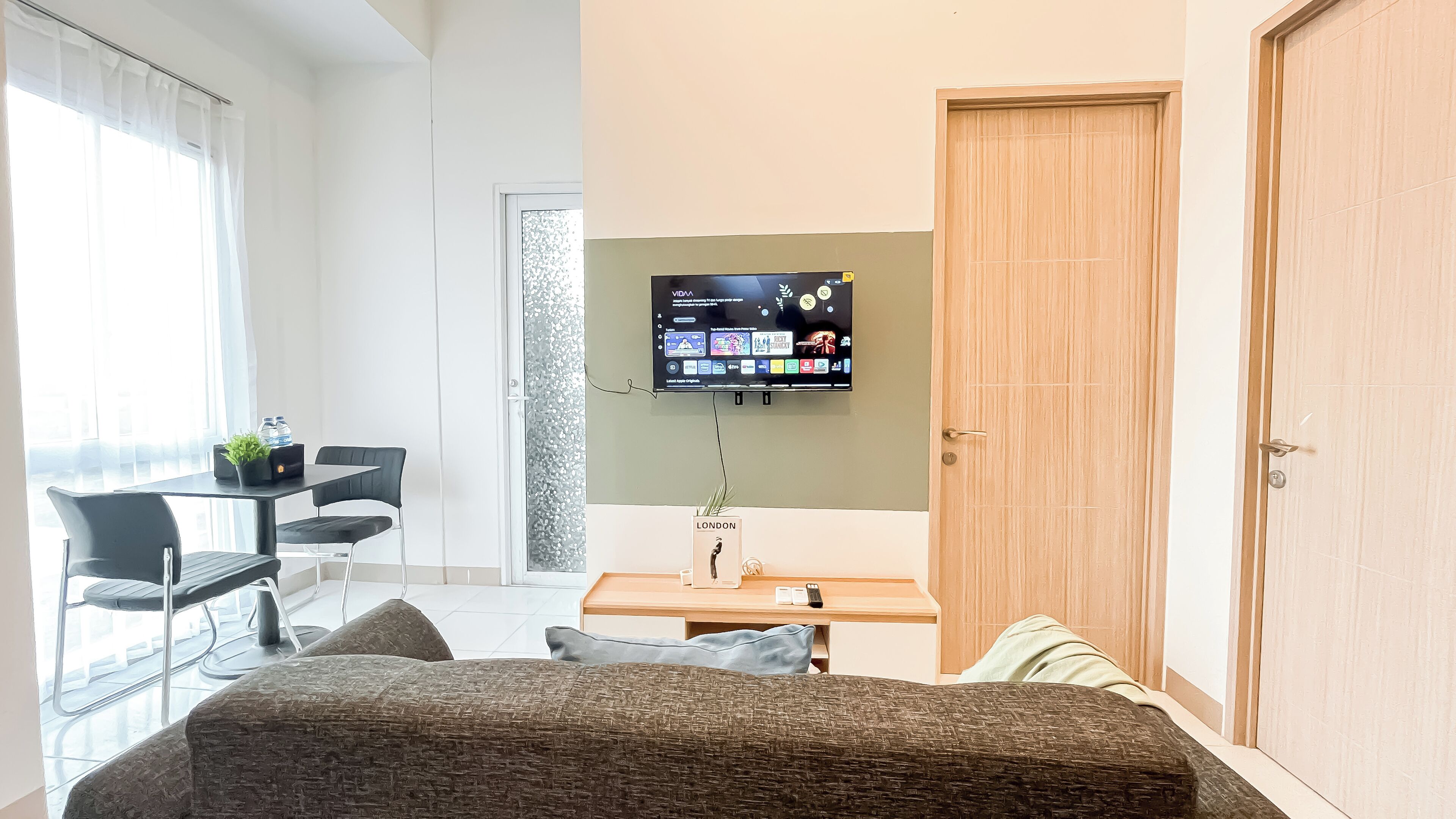 A modern room with a flat-screen TV mounted on the wall, a wooden door, a desk with a plant, and a seating area with a sofa and chairs.