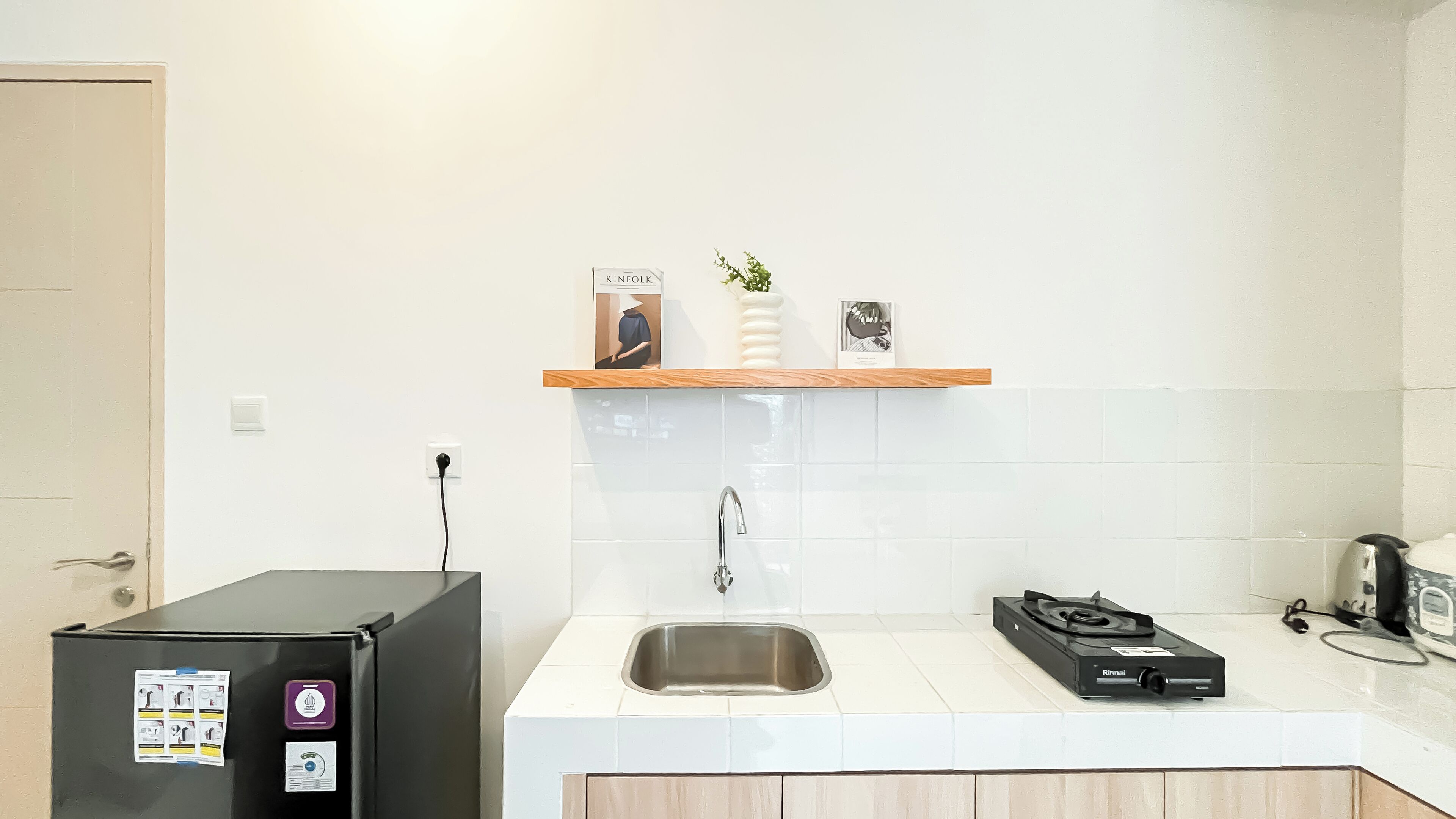 A small kitchenette with a black refrigerator, a sink, a wooden shelf with a plant, and a Rinnai gas stove.