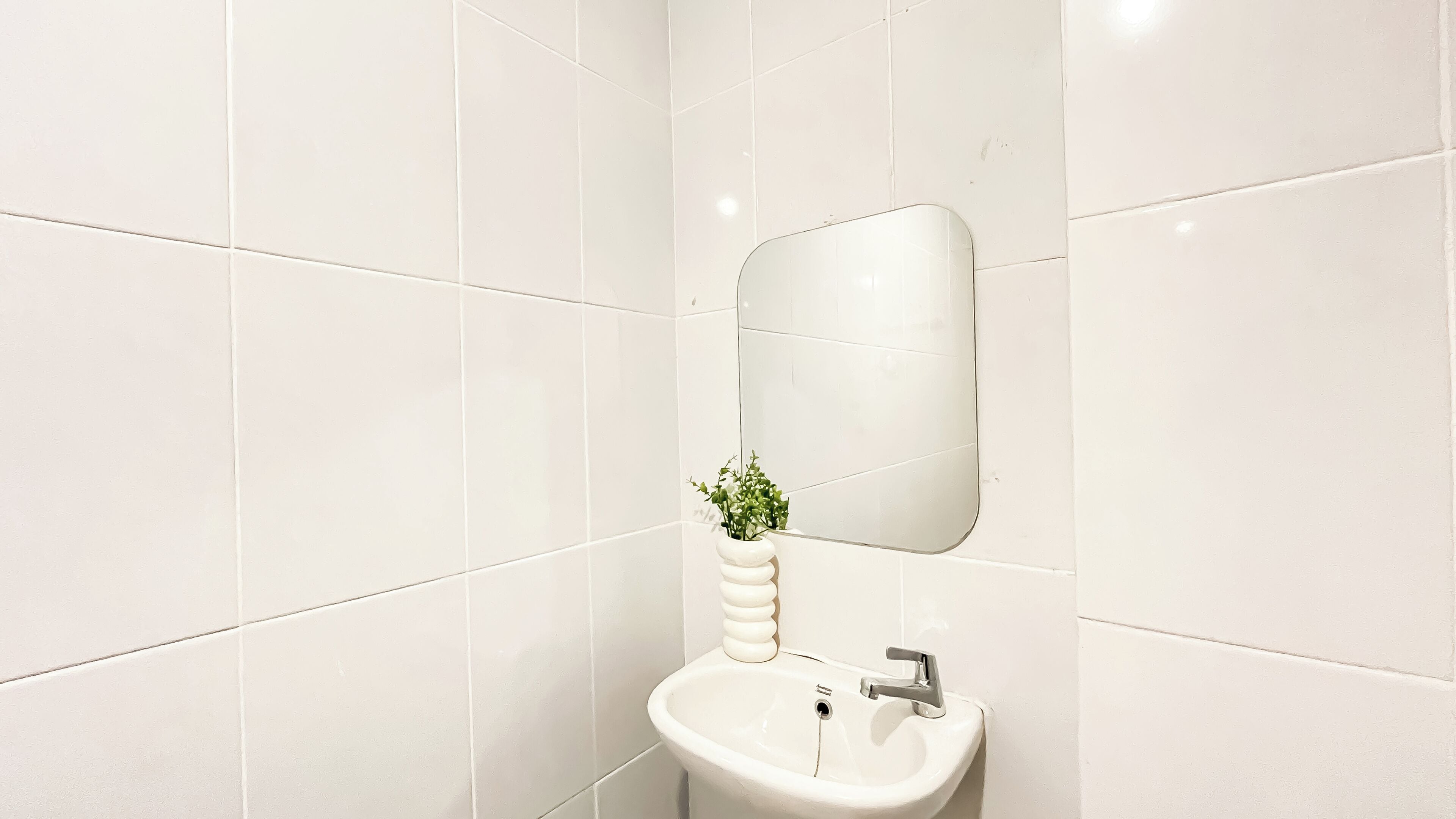 A bathroom with white tiles, a sink, a mirror, and a plant in a white vase.