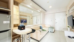 A modern hotel room with a kitchenette, a large mirror, a flat-screen TV, a sofa with cushions, and a small table with a water bottle. - Warm And Comfy 2Br At Green Bay Pluit Apartment (Jakarta)