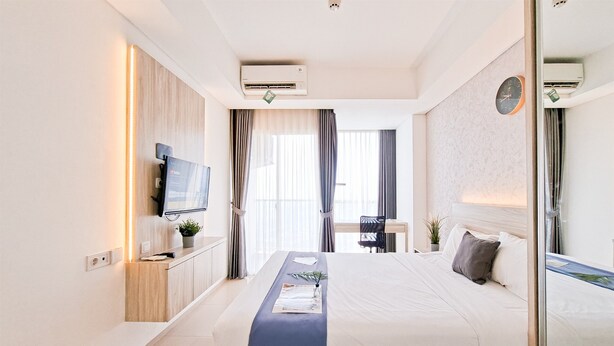 A hotel room with a bed, bedside table, TV, air conditioning unit, and a window with curtains. - Delightfully Cozy Studio Apartment Pacific Garden - Campus Town @Alam Sutera (Tangerang)