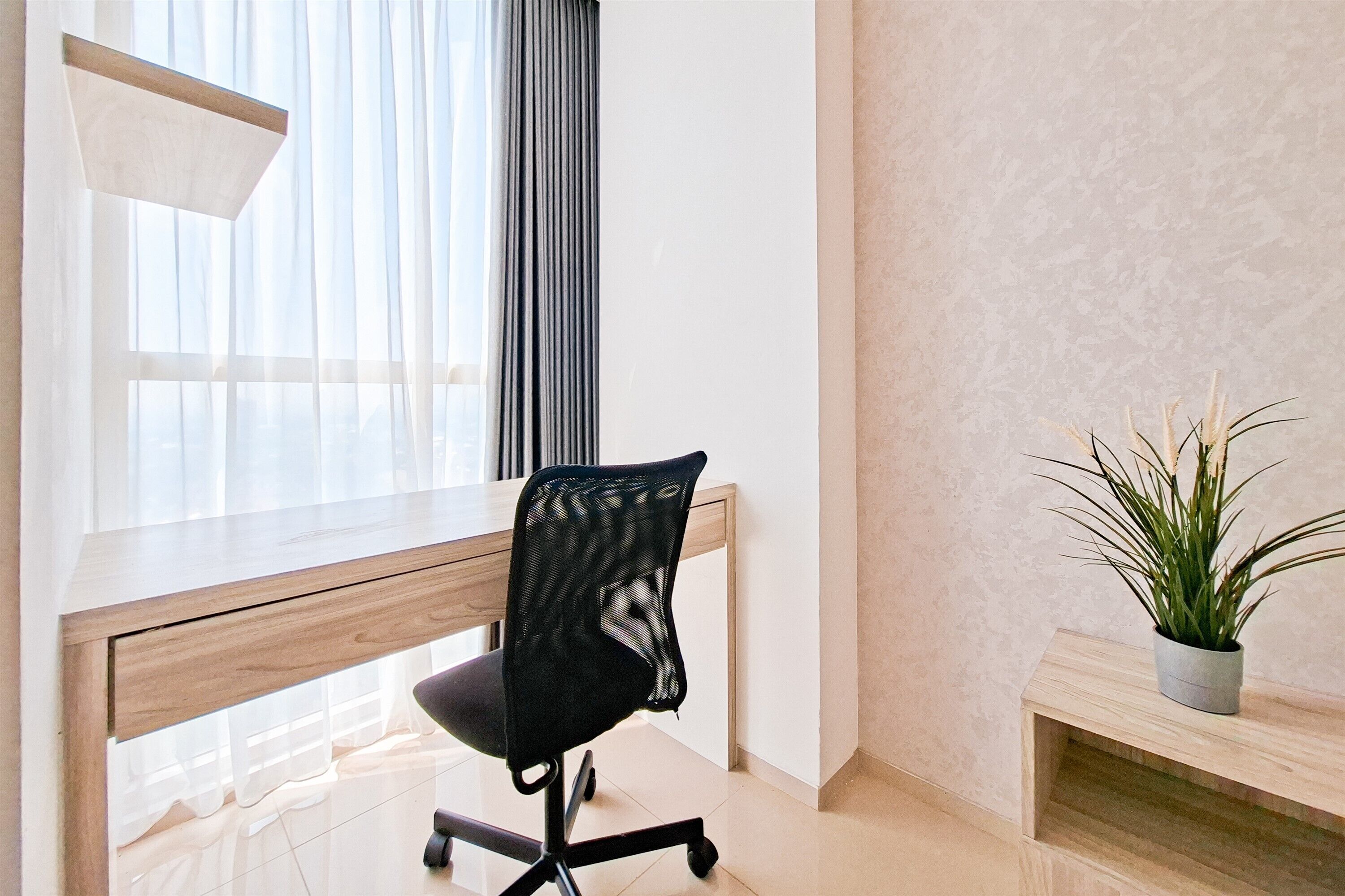 A modern office space with a wooden desk, a black mesh office chair, a potted plant, and sheer curtains allowing natural light.