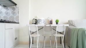 Une cuisine compacte équipée d’une cuisinière à gaz, d’une bouilloire, d’une machine à café et d’une petite plante en pot. On y trouve deux chaises blanches et un canapé recouvert d’un plaid vert.