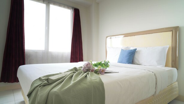 Une chambre d’hôtel avec un lit, une fenêtre avec des rideaux et une table de chevet sur laquelle se trouvent un livre et des fleurs.