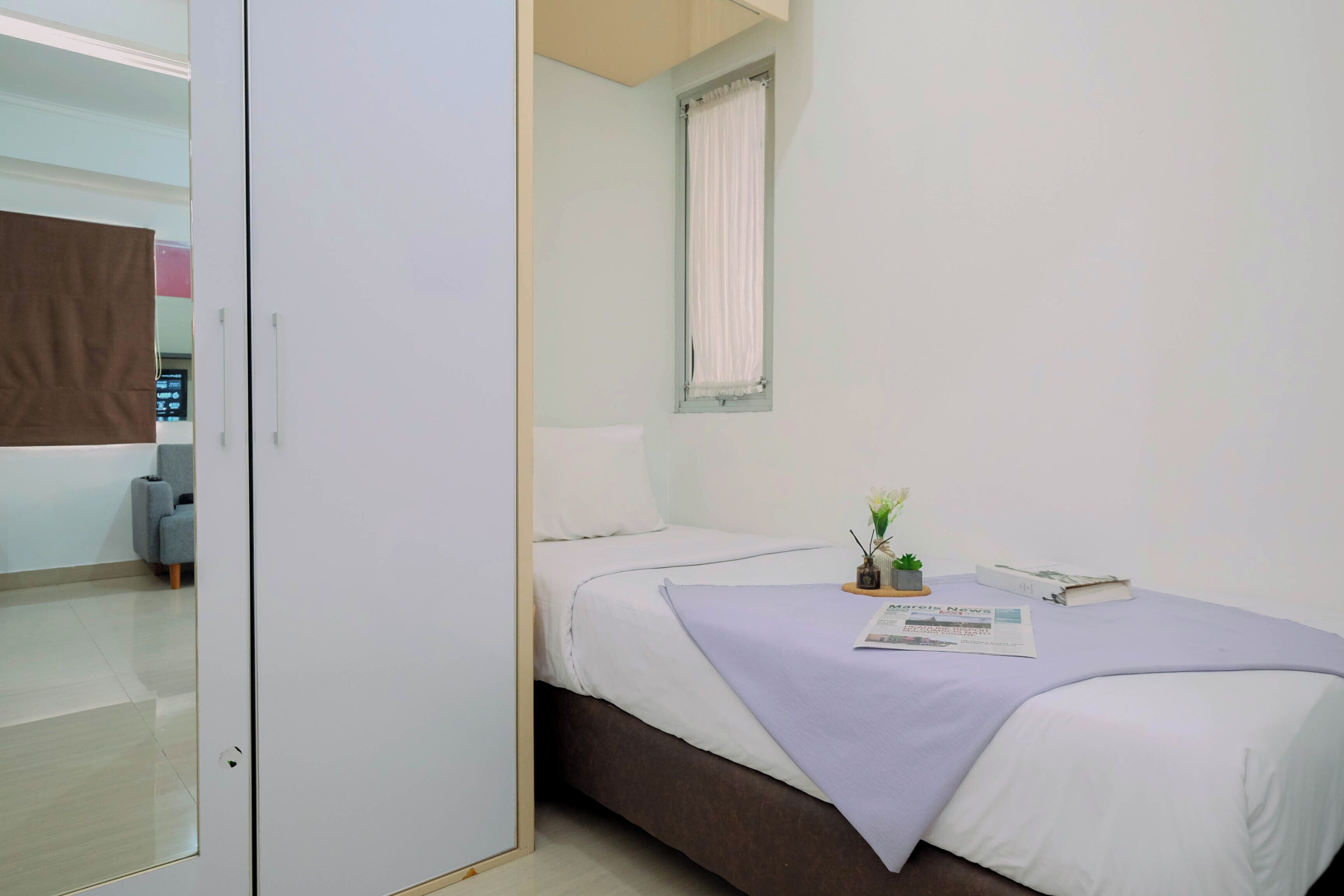 A bedroom with a bed, a window with curtains, a small table with a plant and a book, and a door leading to another room.
