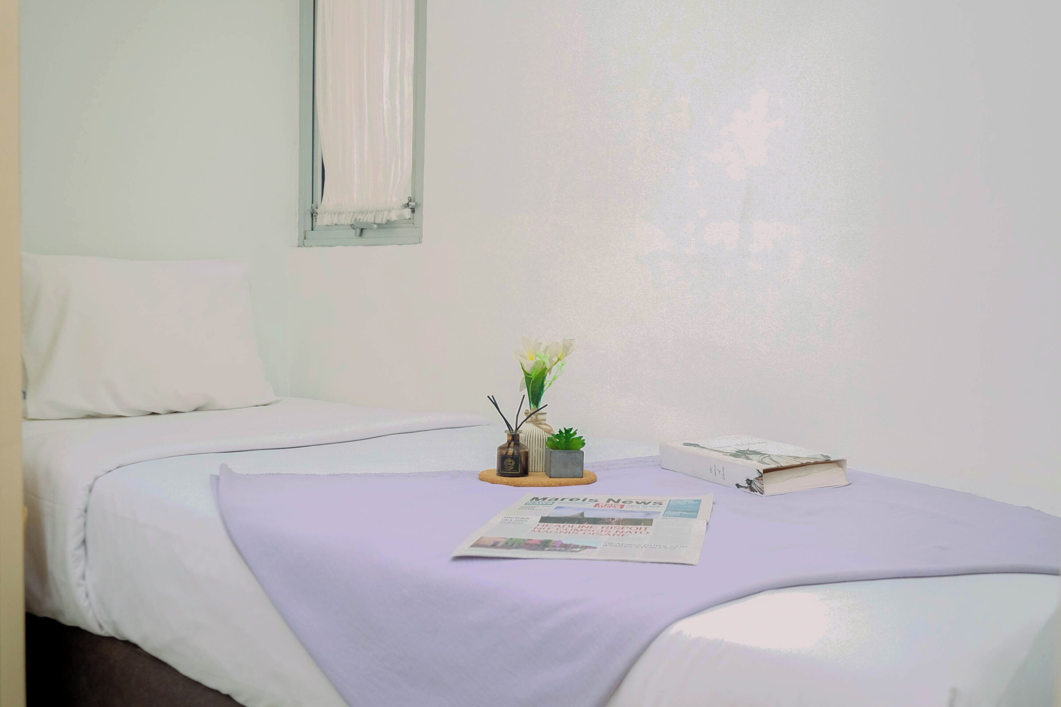 A neatly made bed with a white pillow and a light purple blanket. A small potted plant and a book are placed on the bed.