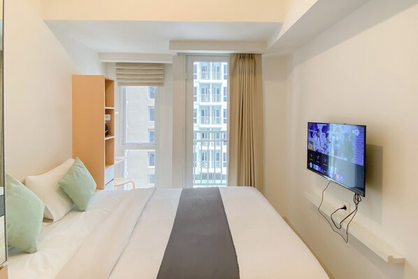 A hotel room with a bed, a television on the wall, a window with curtains, and a view of buildings outside. - Restful Living Studio At Apartment Tokyo Riverside Pik 2 (Teluknaga)
