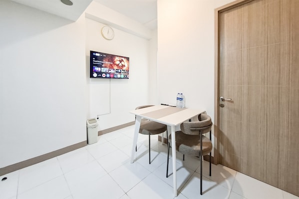 A small, modern room with a white tiled floor, a white table with two chairs, a television mounted on the wall, a door, and a trash bin. - Lovely And Superb 2Br Apartment Tokyo Riverside Pik 2 (Teluknaga)
