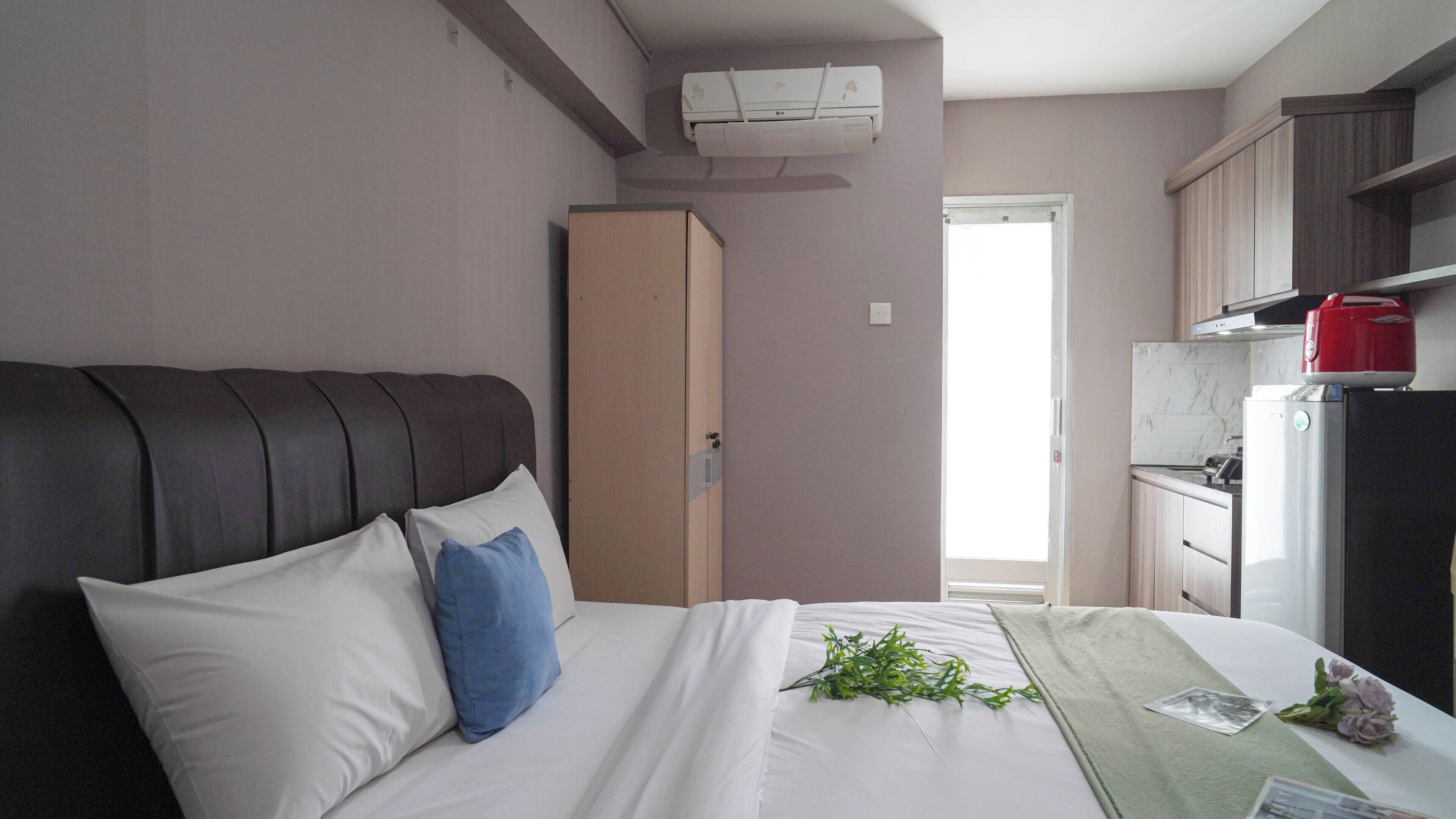 A bedroom with a bed, pillows, a bedside table with a plant, a wardrobe, and a kitchenette with a refrigerator and stove.
