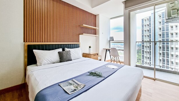 A modern hotel room with a large bed, wooden headboard, and a balcony with a city view. - Enjoyable Living Studio At Apartment Pacific Garden - Campus Town @Alam Sutera (Tangerang)