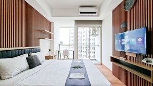 A modern hotel room with a large bed, a flat-screen TV, wooden paneling, and a balcony with a city view. - Enjoyable Living Studio At Apartment Pacific Garden - Campus Town @Alam Sutera (Tangerang)