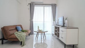 A modern living room with a brown sofa, a glass coffee table with flowers, a white cabinet, and a TV on a stand. - 2Br Near Prasetiya Mulya University At Sky House Bsd Apartment (Jakarta)