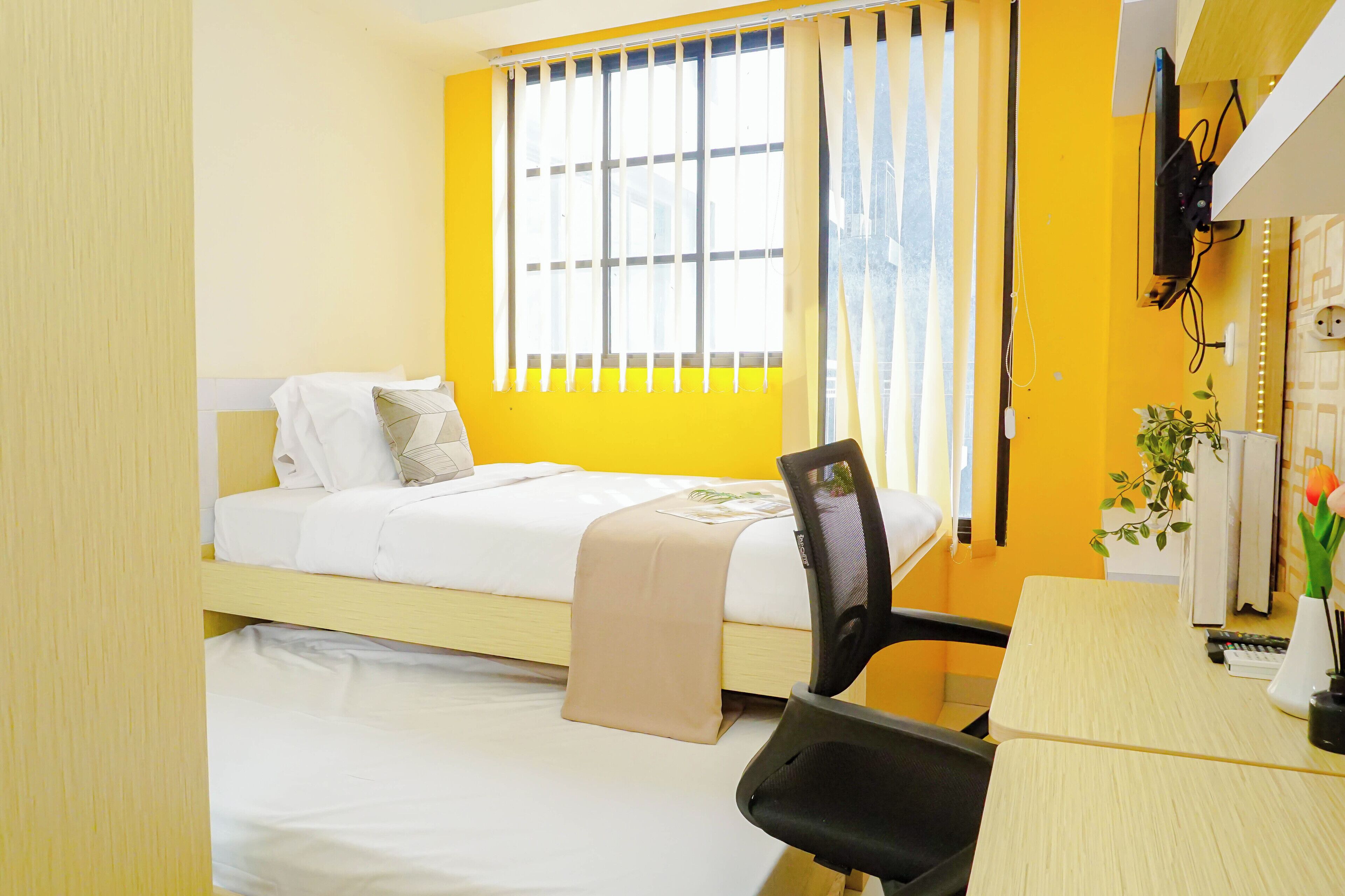 Ein Hotelzimmer mit einem Einzelbett, einem Schreibtisch, einem Stuhl, einem an der Wand befestigten Fernseher und einem Fenster mit Vorhängen.