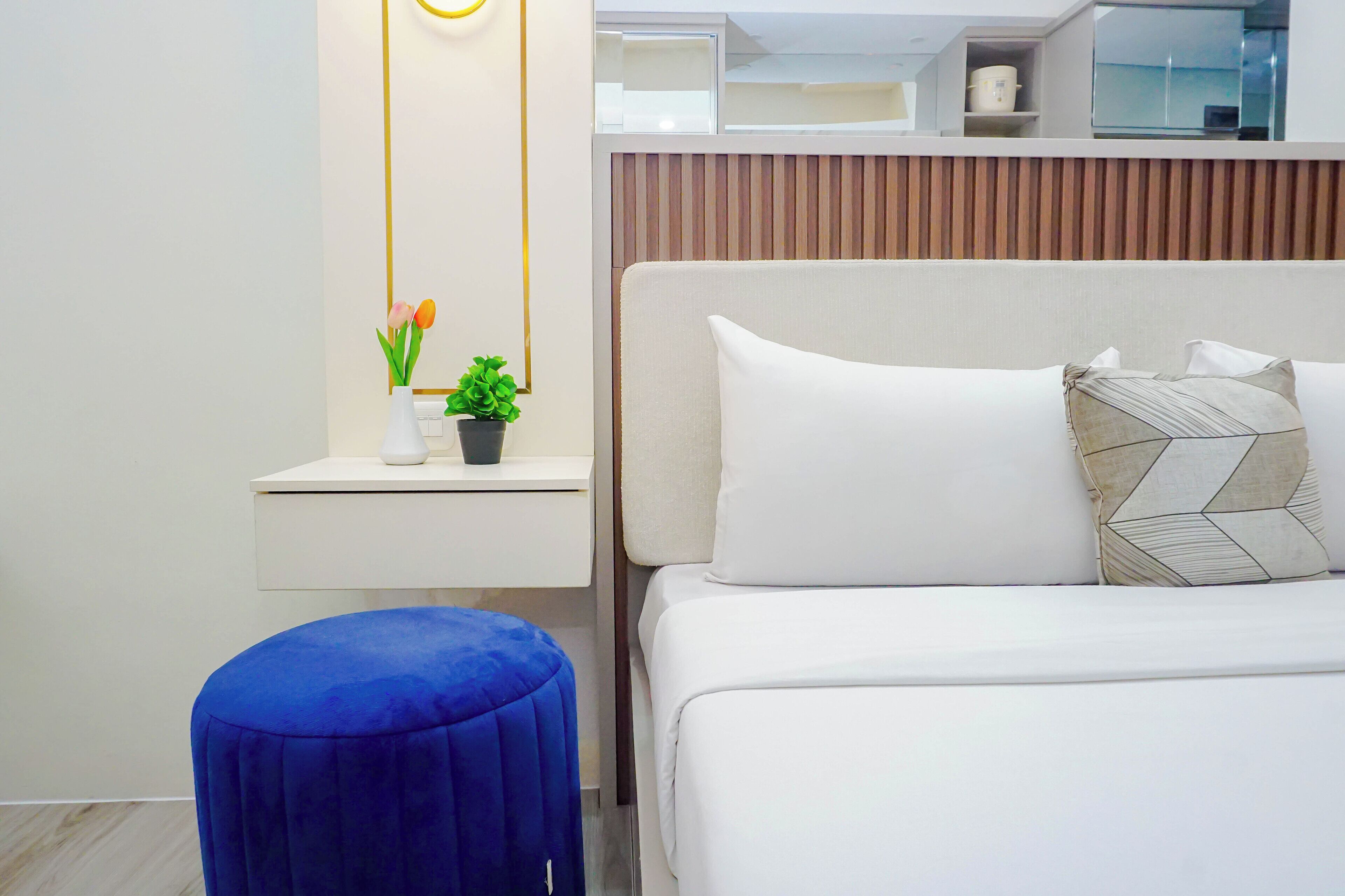 A modern hotel room with a bed, a small desk with a plant, and a blue ottoman.
