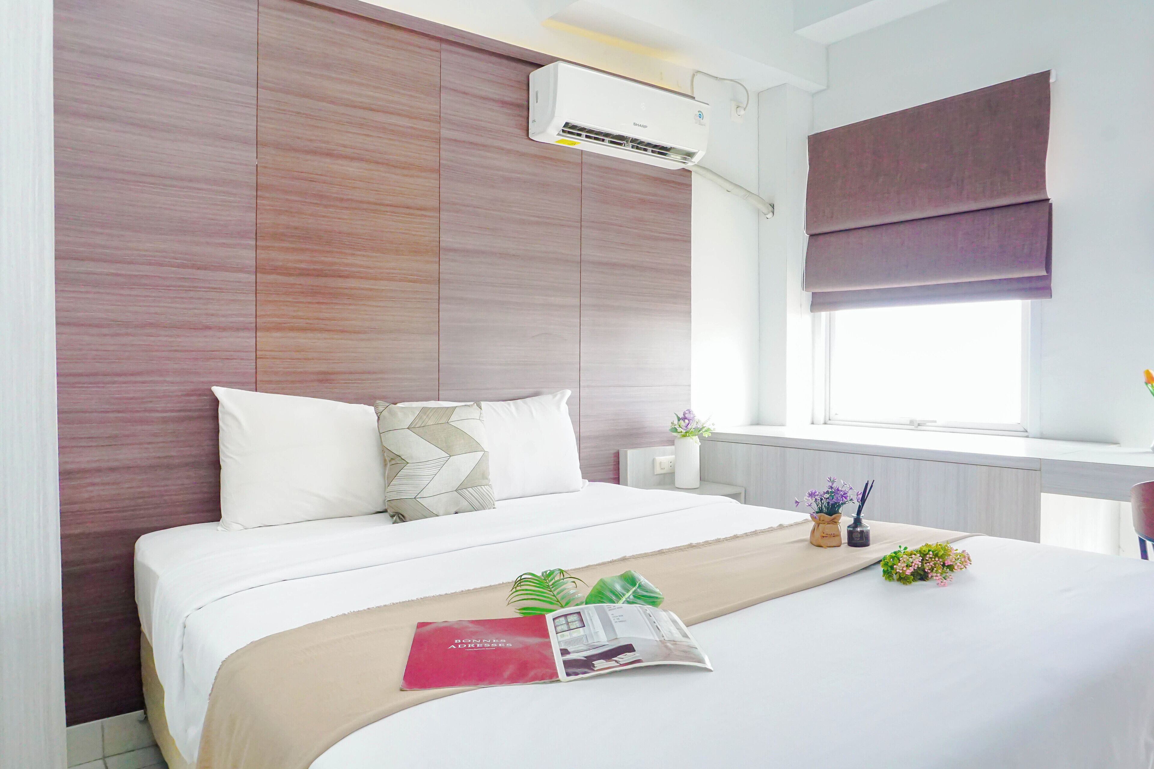 Une chambre d’hôtel avec un lit, une fenêtre avec un store, une table de chevet avec un vase et un magazine, et un appareil de climatisation fixé au mur.