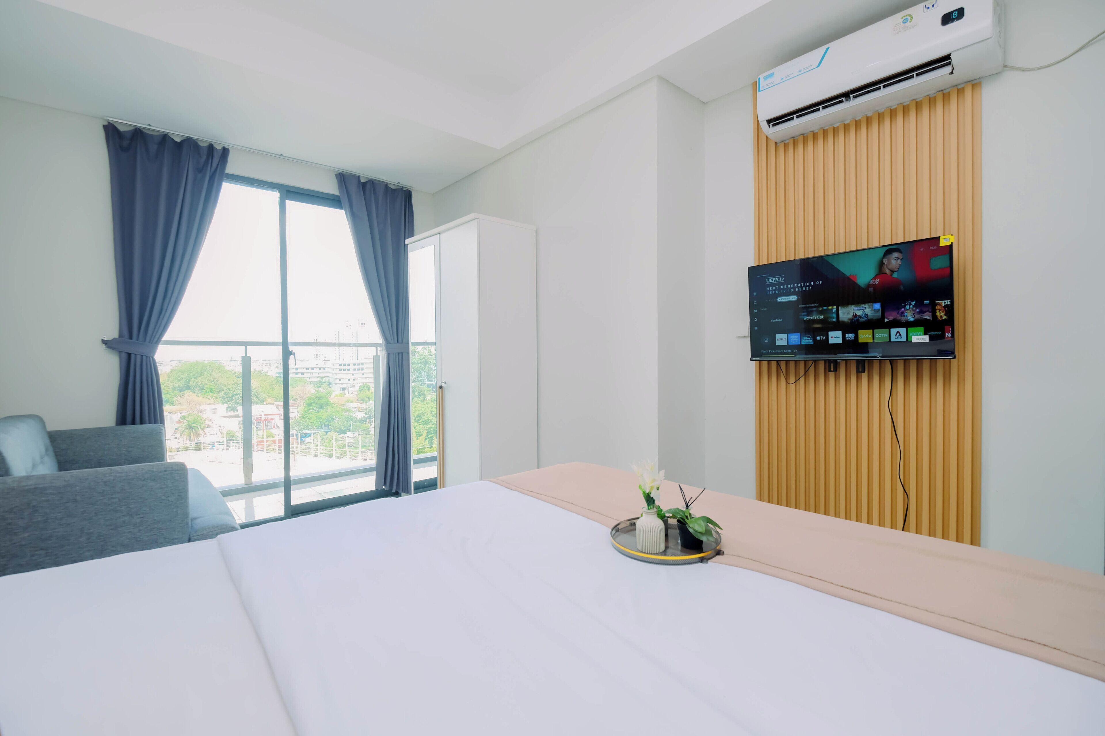 A hotel room with a bed, a TV mounted on a wooden panel, a window with curtains, and a view of the city.