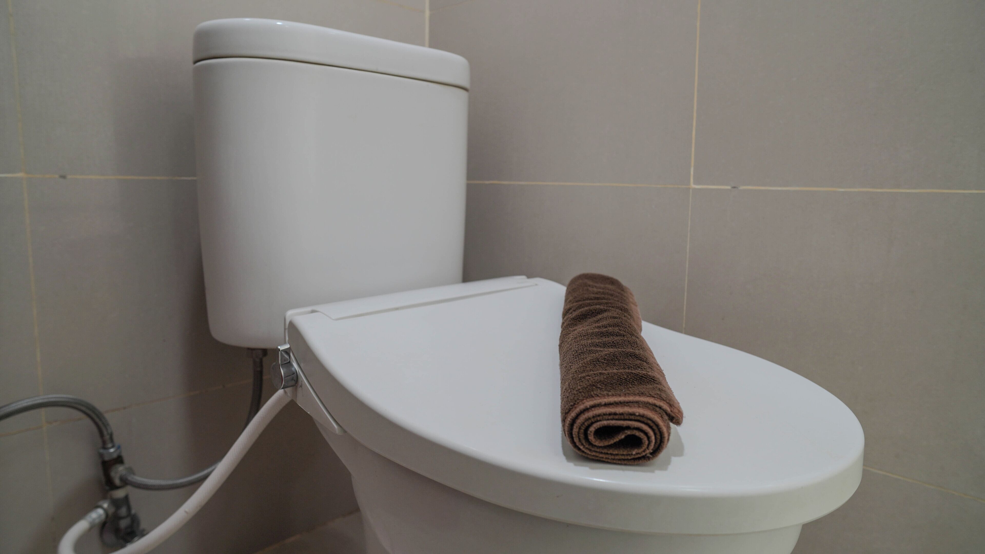 Et hvidt toilet med et brunt håndklæde på sædet, en synlig skyllearmatur og et synligt rør.