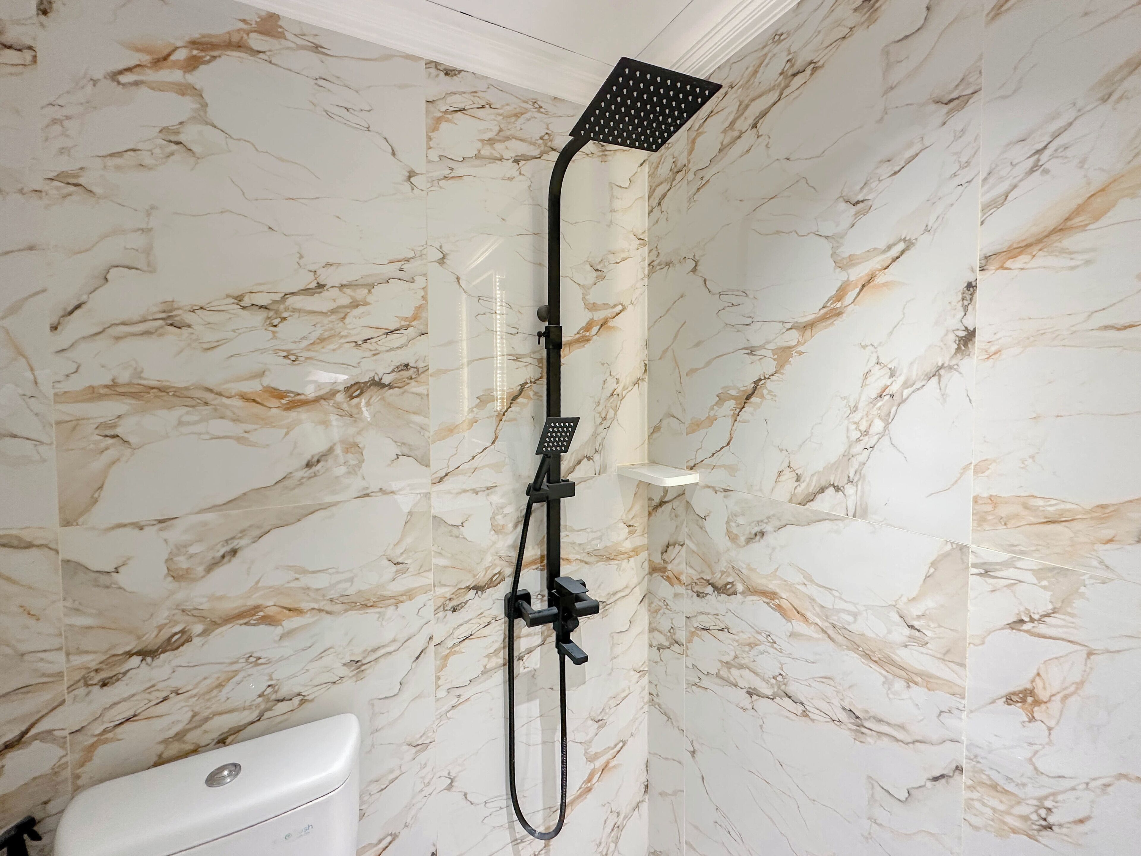 Une salle de bain avec un mur en marbre, une douche équipée d’un pommeau de douche à main et des toilettes blanches.
