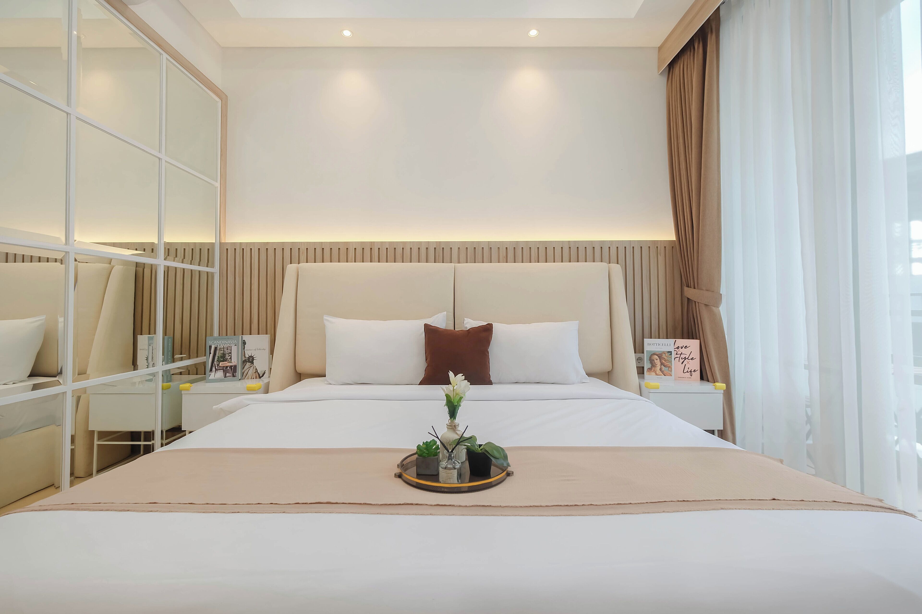 Una habitación de hotel moderna con una cama grande, una mesita de noche con una bandeja de plantas y otra con un libro, y un espejo amplio.
