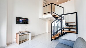 A modern living room with a flat-screen TV mounted on the wall, a wooden desk, a grey sofa, and a black spiral staircase. - Cozy White Loft Studio At Kingland Avenue Apartment (South Tangerang)