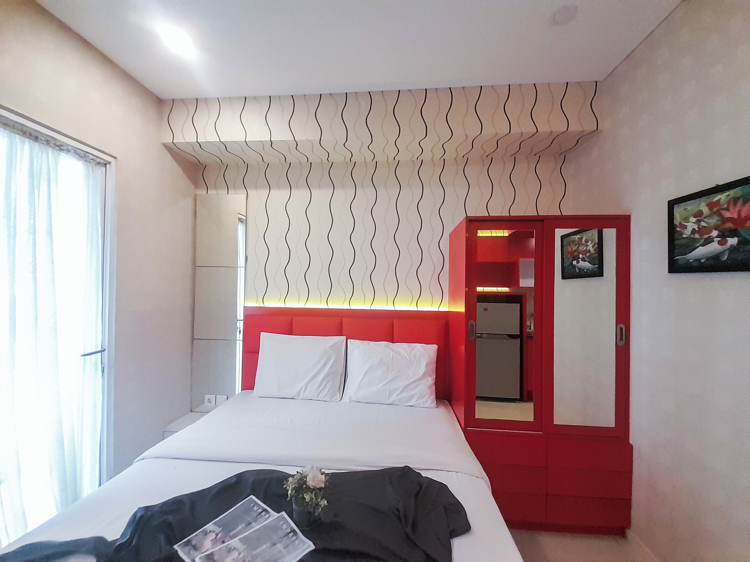 A bedroom with a bed, a red wardrobe, a mirror, and a window with curtains.
