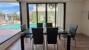 Un coin repas avec une table et des chaises en verre, de grandes fenêtres donnant sur une piscine et sur la verdure.