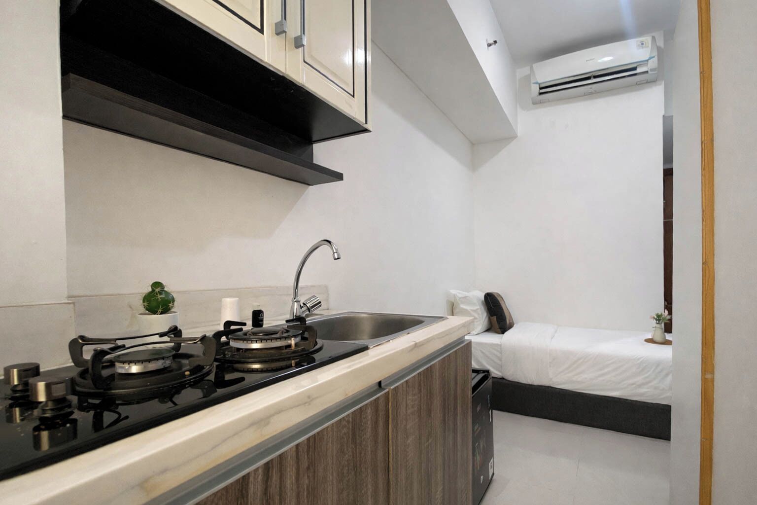 A compact kitchen with a gas stove, sink, and a small bedside table with a pillow and a plant.