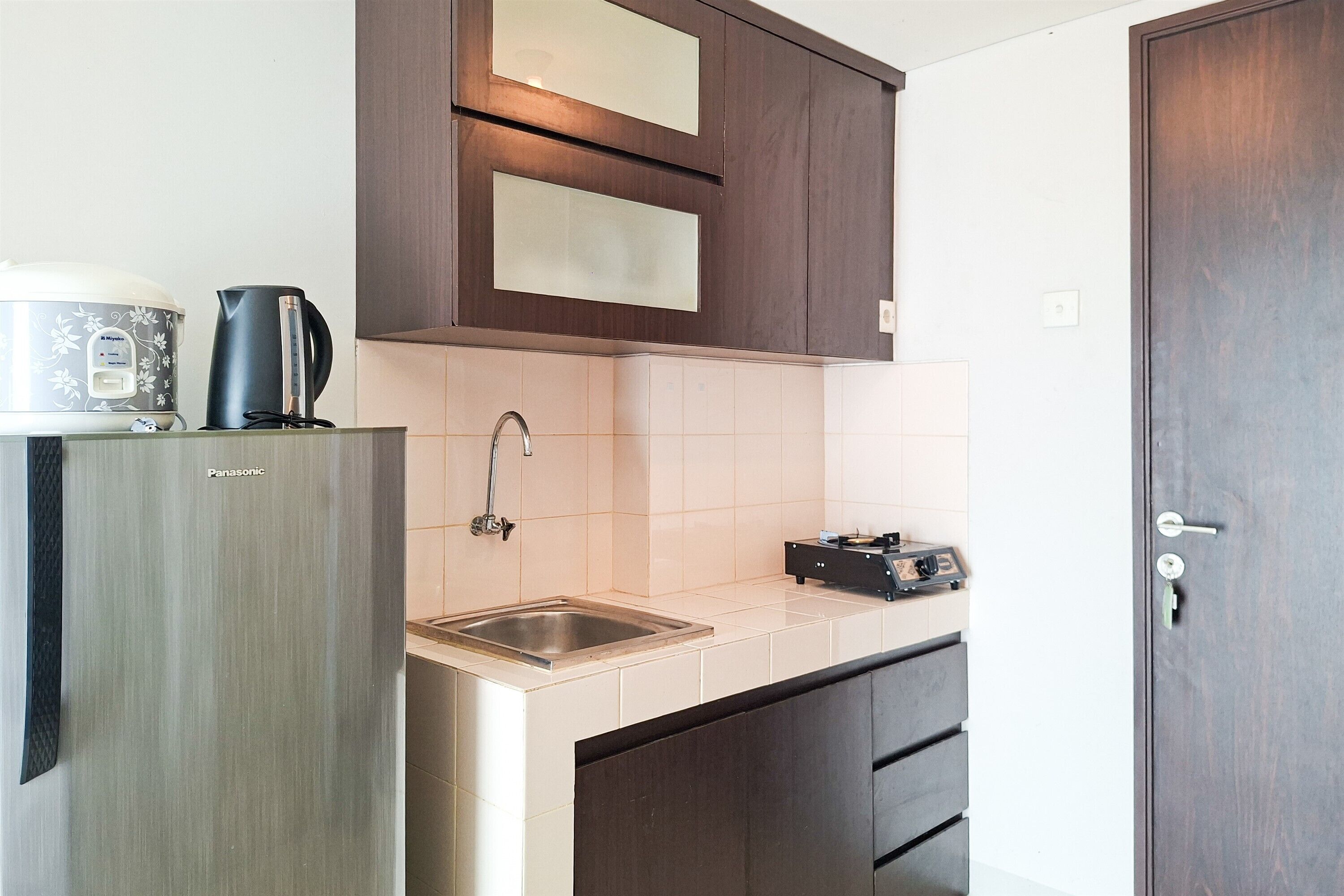 Una cocina compacta con un lavamanos de acero inoxidable, una tetera y una cocedora de arroz sobre la encimera.