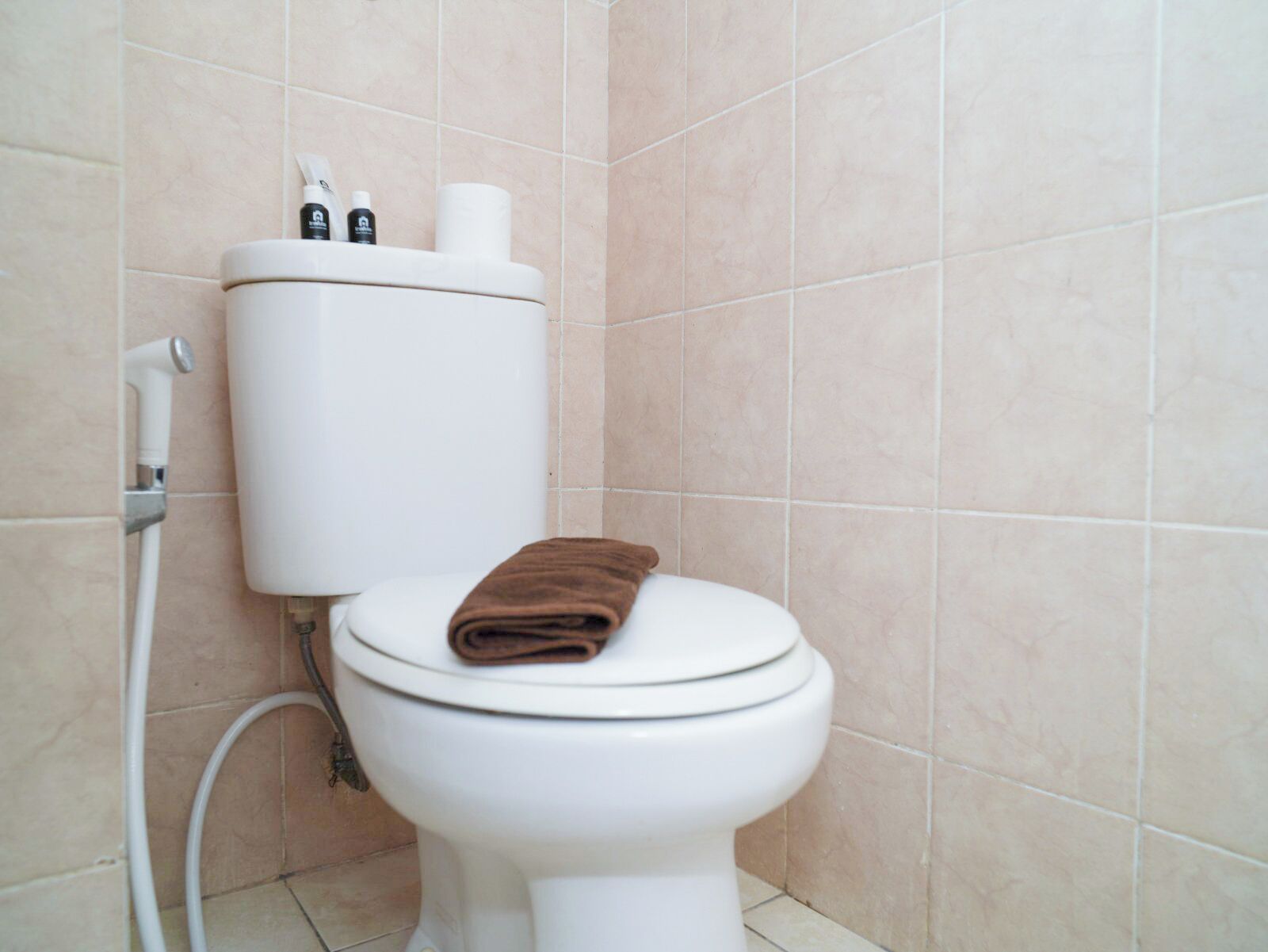 A white toilet with a brown towel on the seat, a toilet paper roll, and a bidet attachment.
