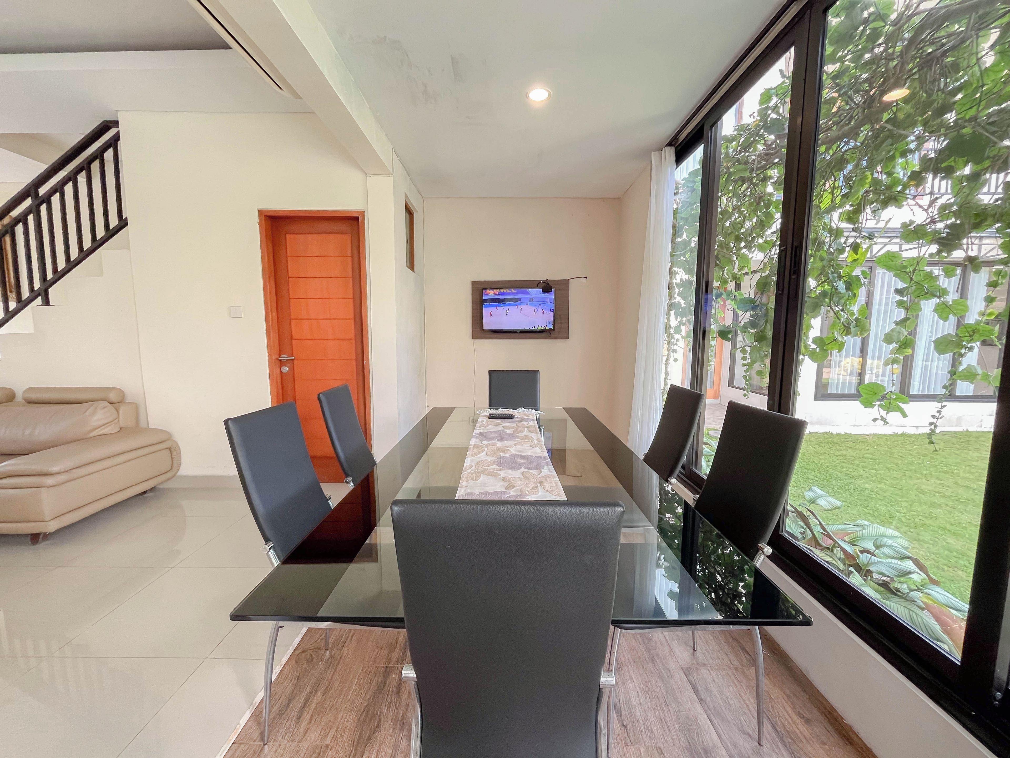 A modern living room with a glass dining table, black chairs, a beige sofa, and a TV on the wall.