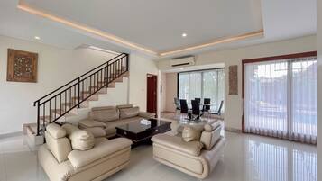 A spacious living room with a beige sectional sofa, a coffee table, a staircase, and a dining area with a glass table and chairs.