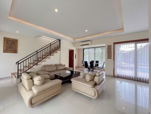 A spacious living room with a beige sectional sofa, a coffee table, a staircase, and a dining area with a glass table and chairs. - 3BR Jimbaran Villa at Casa De Uli Bali (Jimbaran)